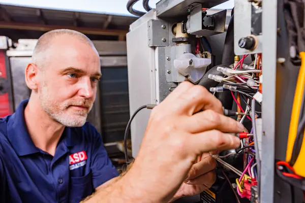 A concentrated male technician with a beard, wearing a dark blue ASD polo shirt, is installing electrical components within a large gray machine. He's manipulating cables and examining the connections closely. The focus is on his hands and the complexity of the task at hand.