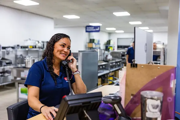 A smiling woman with curly hair, wearing a blue polo shirt with an ASD logo, is talking on a telephone at a customer service desk.