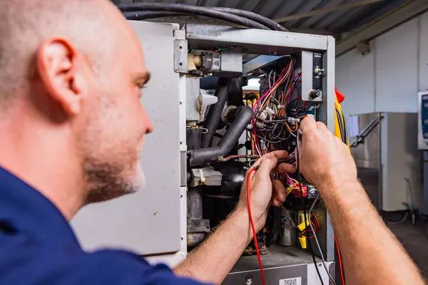 A focused ASD technician in a dark blue shirt is working on an intricate piece of machinery, using a multimeter on its electrical components. His attention is directed towards the tangle of wires and tubes inside the open panel of the equipment.