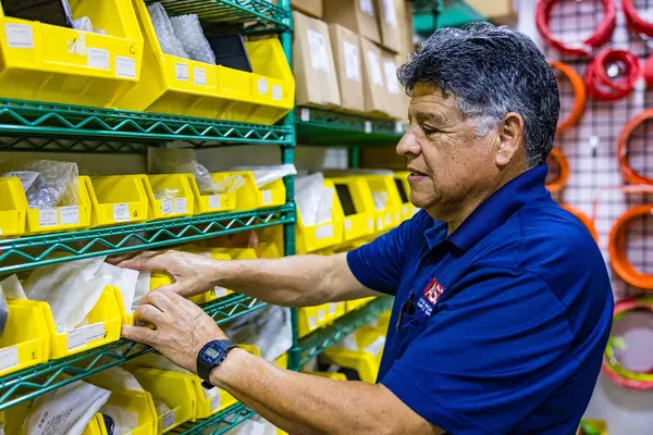 An employee at ASD's Parts Center, a man with gray hair, is selecting parts from a shelf stocked with yellow bins labeled with part numbers. He is wearing a dark blue polo shirt with the ASD logo and is examining an item in his hand. The background shows an organized warehouse environment with various parts and tools on shelving units.