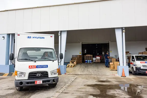 A ASD delivery truck is parked outside a warehouse loading dock. The warehouse door is open, revealing pallets of boxed goods and a person standing inside. Another white ASD van is parked to the right.